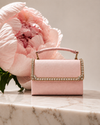 Blush pink satin mini top handle bag with crystal embellished flap edge, placed on marble surface beside pink peony bloom, The House of Shine Bijou Edit