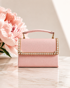 Blush pink satin mini top handle bag with crystal embellished flap edge, placed on marble surface beside pink peony bloom, The House of Shine Bijou Edit