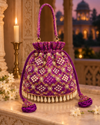 Purple Gulnaar Bandhej potli with gold embroidery and pearl-drop edging beside a candle at dusk.