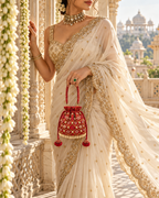 Model in an ivory and gold saree carrying the red Gulnaar potli beside a carved palace doorway.