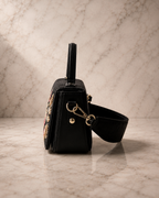 Side view of The House of Shine Florence hand-embroidered black satchel showing detachable crossbody strap and gold hardware on marble surface
