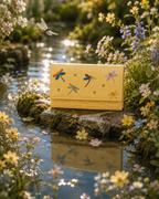 Libellula butter-yellow hand-embroidered dragonfly mini bag by The House of Shine, photographed beside a flower-filled garden pond.