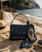 Mirtillo embroidered crossbody bag resting on sand with a silk scarf, straw hat, and fresh blueberries