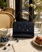Mirtillo luxury embroidered satchel on a café table with cappuccino, croissant, and fresh blueberries