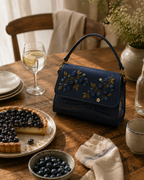 Mirtillo luxury embroidered satchel on a rustic wooden table beside a blueberry tart and glass of citrus water