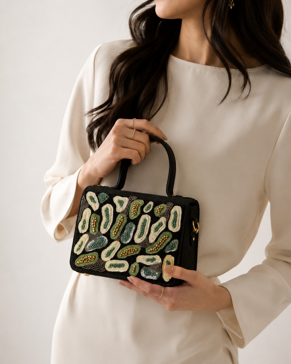 Model holding The House of Shine Prisma hand-embroidered black satchel with green and ivory sequin and bead motifs, styled with a cream dress