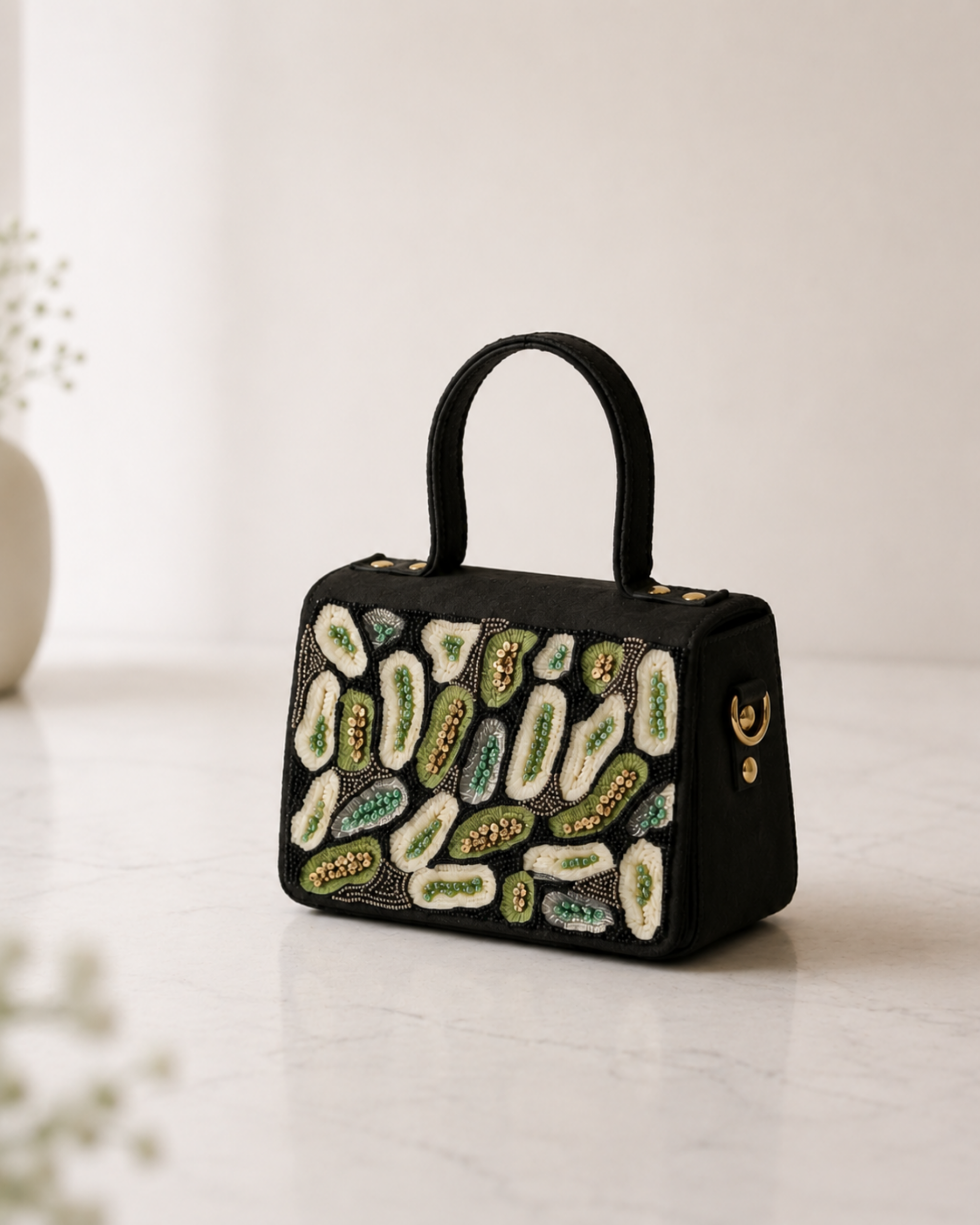 The House of Shine Prisma hand-embroidered black satchel with green and ivory sequin and bead motifs, angled view on marble surface with soft botanical background