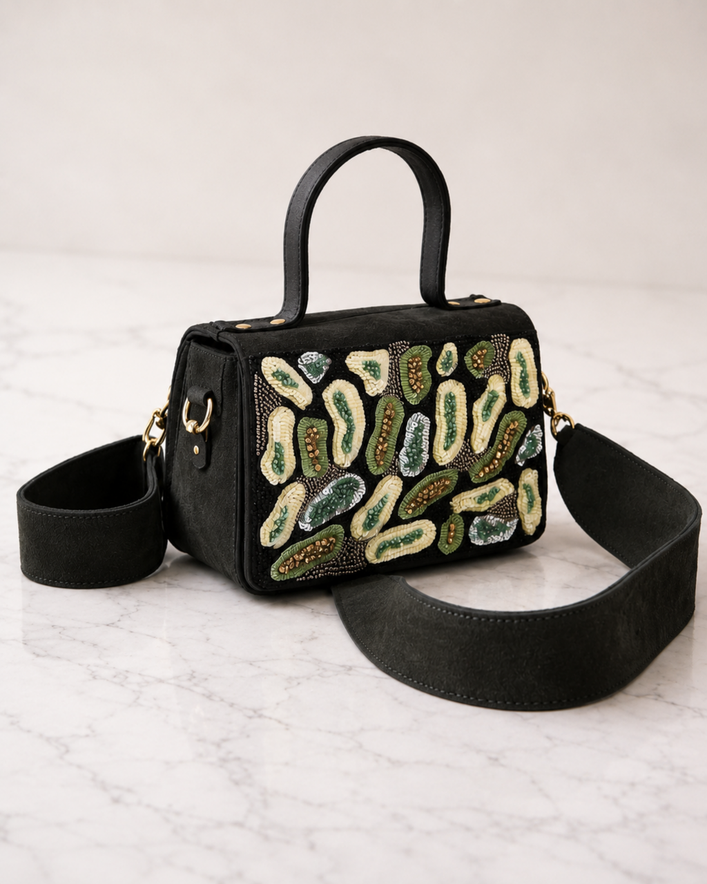 The House of Shine Prisma hand-embroidered black satchel with green and ivory sequin and bead motifs, shown with detachable wide black crossbody strap on marble surface
