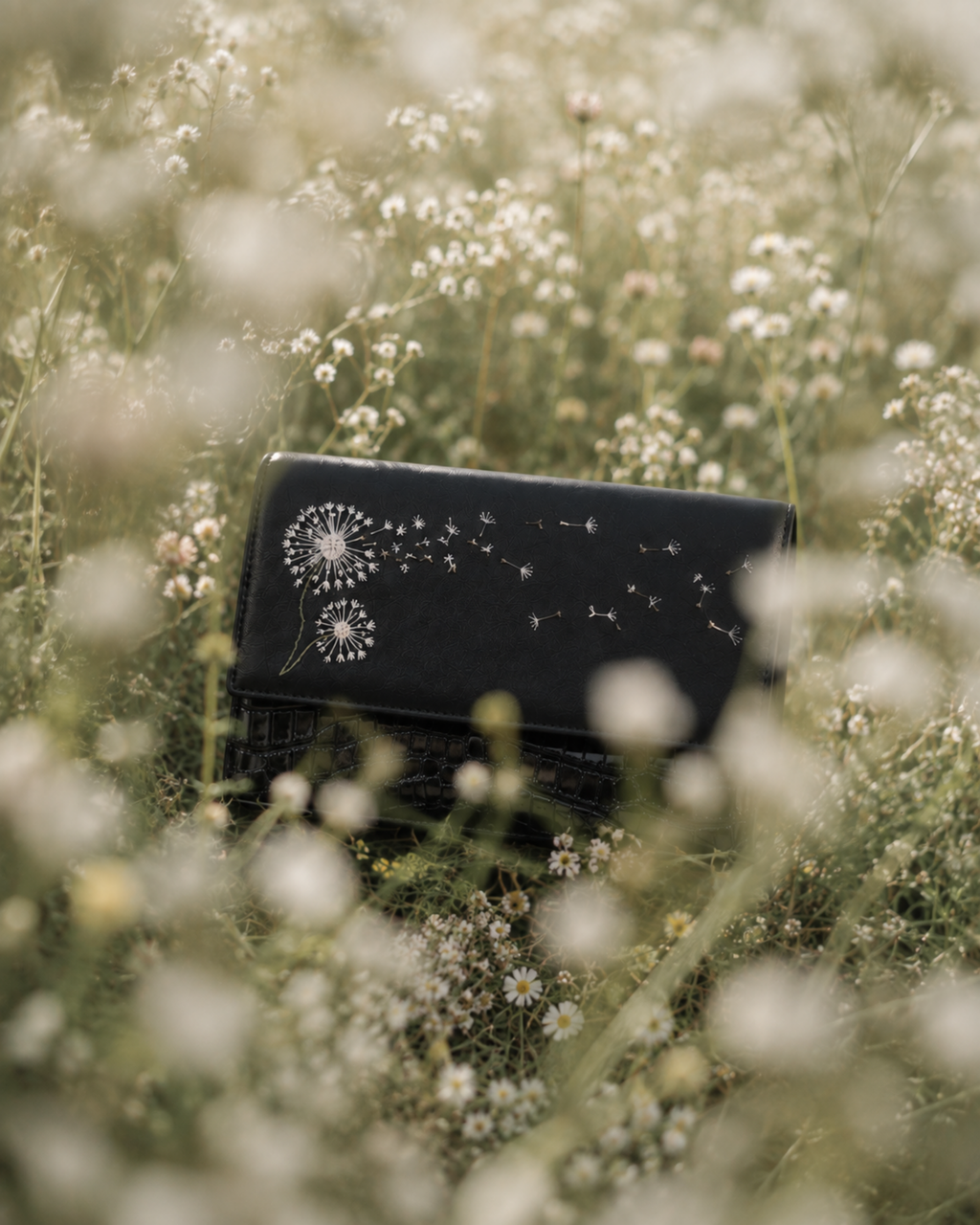 Soffio black mini bag with white dandelion embroidery photographed among soft wildflowers in a dreamy meadow.
