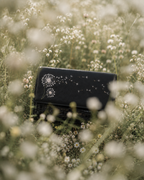Soffio black mini bag with white dandelion embroidery photographed among soft wildflowers in a dreamy meadow.