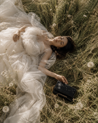 Woman in an ivory dress lying in a meadow beside the Soffio black dandelion embroidered mini bag.