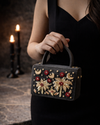 Model holding House of Shine Tropical Moonbeam Satchel in charcoal velvet wearing a black dress, gold and red zardozi embroidered luxury handbag styled for evening