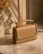 Liquid Gold crystal-embellished mini top-handle bag by House of Shine, angled detail shot on marble with dried golden botanicals, gold-polished hardware, saffiano exterior, Bijou Edit.