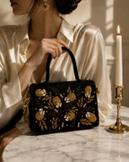  Nocturne Satchel held by top handle by woman in cream silk with gold earrings beside brass candlestick, Noir Atelier by The House of Shine