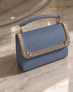 Something Blue crystal-embellished mini top-handle bag by House of Shine, angled detail shot on sunlit marble showing hand-set crystal trim and saffiano texture, gold-polished hardware, Bijou Edit.